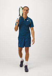 Jūros karininkų sportinė polo marškiniai su žaliais akcentais, derančios tamsiai mėlynos šortai ir tamsūs sportbačiai. Laiko teniso raketę, turi struktūrizuotą audinį.