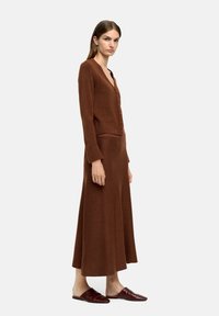 Brązowy, dzianinowy długi kardigan z dekoltem w serek i detalami guzikowymi, w zestawie z dopasowaną długą spódnicą. Gładka tekstura, dopasowany krój, bezszwowa konstrukcja.