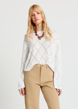 Pull en maille blanche avec motifs en losange, manches larges, associé à un pantalon beige taille haute. Collier de perles vertes et rouges en accent.