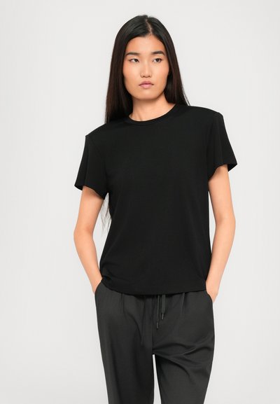 Young woman with long straight hair wearing a black short-sleeve shirt and dark gray pants, standing with hands in pockets against plain background.