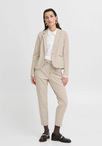 Béžový blazer se jednoduchým designem v kombinaci s úzkými kalhotami ve stejné barvě. Bílé tričko pod ním, nošené s tmavými mokasíny a vzorovanými ponožkami.