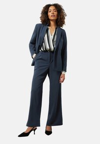 Marineblauw kostuum met wijde broekspijpen en een op maat gemaakte blazer, gecombineerd met een gestreepte blouse. De schoenen zijn zwart en hebben een slank, puntig ontwerp.