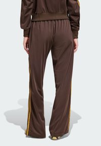 Pantaloni atletici din satin marron cu dungi galbene pe laterale, având un elastic în talie și un design cu picioare evazate. Textură netedă și croială lejeră.