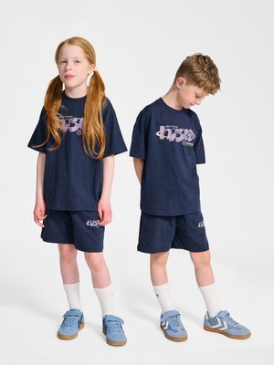 Dwoje dzieci ubranych w pasujące granatowe stroje sportowe z jasnofioletowymi logo, białe skarpetki i jasnoniebieskie trampki, stojących na jednolitym tle.