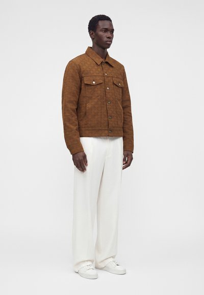 Homme debout portant une veste marron texturée, un pantalon blanc à coupe large et des baskets blanches sur un fond clair uni.