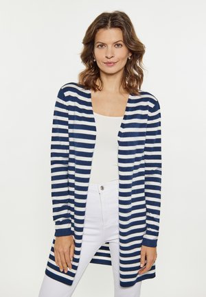 Frau trägt einen langen marineblau-weiß gestreiften Cardigan, ein weißes Top und weiße Hosen und steht vor einem schlichten hellen Hintergrund.