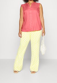 Jasnoróżowy top bez rękawów z koronkowym wykończeniem, w połączeniu z szerokimi żółtymi spodniami w serca. Modelka trzyma białą torebkę.