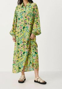 Robe florale longue verte avec des motifs roses et bleus, boutonnée sur le devant et avec des manches larges. Associée à des sandales beige clair avec des semelles noires.