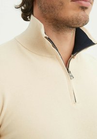 Pull beige en tricot avec un col montant et une fermeture éclair quart. Doté d'une doublure bleu foncé à l'intérieur du col. Texture douce.