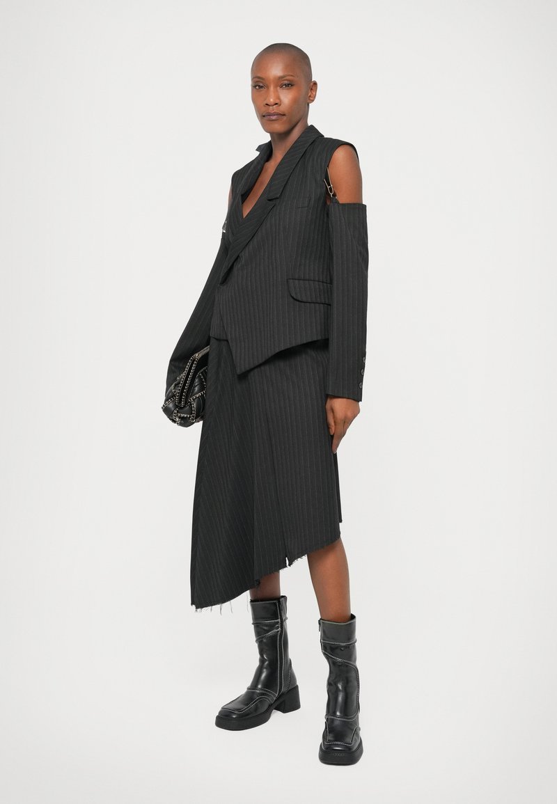 Maßgeschneiderter schwarzer Nadelstreifenanzug mit asymmetrischem Rock und ärmellosem Blazer, kombiniert mit kniehohen schwarzen Stiefeln.