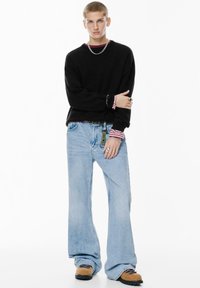 Maglione nero con polsini a coste, jeans a gamba larga blu chiaro e zoccoli color beige. Gli accessori includono una collana a catena in argento e anelli.