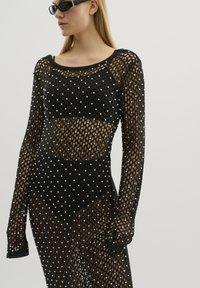 Rochie din plasă neagră, cu o croială lejeră, având accente din perle pe întreaga suprafață, mâneci lungi și decolteu rotund. Design texturat, transparent.