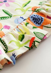 Bomuldsstof med et farverigt blomstermønster, der har blå fugle, grønne blade og orange, pink og gule blomster. Volangdetalje ved kanten.