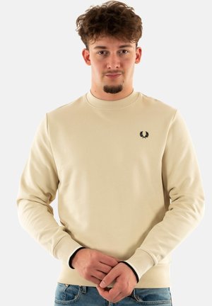 Sudadera beige con mangas largas, cuello redondo, puños acanalados y un pequeño logo negro bordado en el pecho. Textura de tela suave.