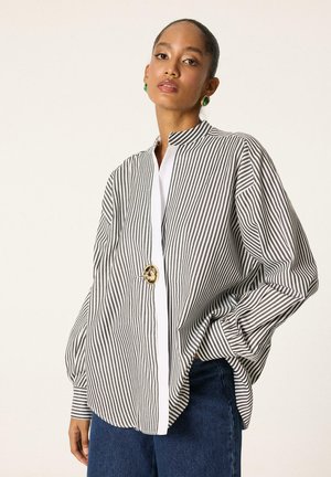 Γυναίκα που φοράει ένα φαρδύ μακρυμάνικο πουκάμισο με ασπρόμαυρες ρίγες και ένα μεγάλο χρυσό κουμπί, συνδυασμένο με μπλε τζιν, στέκεται μπροστά από ένα απλό φόντο.