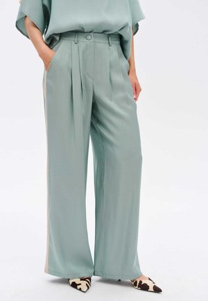 Pantalons larges vert menthe en tissu lisse, avec des plis et une taille haute. Associés à des chaussures à bout pointu en imprimé vache.