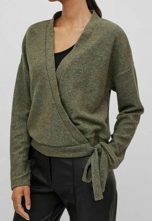 Donna che indossa un maglione verde avvolgente legato in vita sopra una maglietta nera con pantaloni neri, in piedi davanti a uno sfondo semplice.