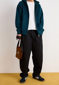 Tealfärgad zip-up hoodie, vit t-shirt, svarta vida byxor och mörka sneakers med vita ränder. Brunt kordflöjls crossbody-väsk.