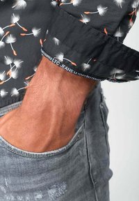 Camisa negra con patrón de dientes de león en blanco y naranja, con los puños enrollados que muestran la etiqueta de marca; combinada con jeans de mezclilla gris desgastados.
