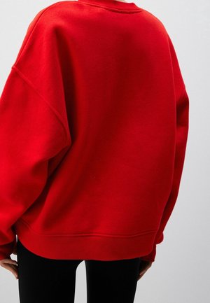 Personne portant un sweat-shirt rouge vif surdimensionné avec ourlet et poignets côtelés, debout devant un fond clair uni.