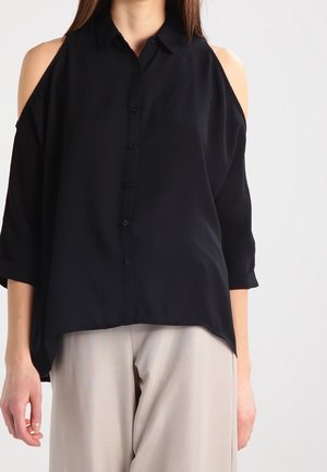 Žena v černé halence s odhalenými rameny a knoflíky, doplněná světlými béžovými volnými kalhotami na neutrálním pozadí.