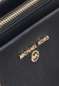 Crni novčanik teksturiranog izgleda s zlatnim patentnim zatvaračem i detaljima. Ima "MICHAEL KORS" logo i okrugli "MK" simbol na prednjoj strani.