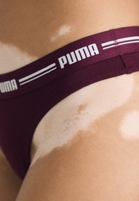 Maroon pamučni tangi s širokim pojasom koji ima bijele pruge i "PUMA" logotip. Bliski pogled koji naglašava teksturu tkanine i kroj.