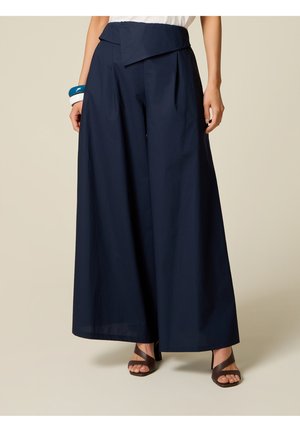 Vrouw draagt marineblauwe wijde broek met asymmetrische tailleband, witte top, bruine sandalen met hak en blauwe en witte armbanden om linkerpols.