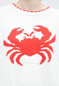 Λευκό πλεκτό πουλόβερ με έντονο κόκκινο σχέδιο καβουριού στο κέντρο του μπροστινού μέρους, με υφή και ραμμένο λαιμόκοψη με βολάν.