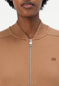 Chaqueta bomber marrón con cremallera, que presenta un cuello y puños acanalados, textura suave, y un pequeño logo cerca de la cremallera.