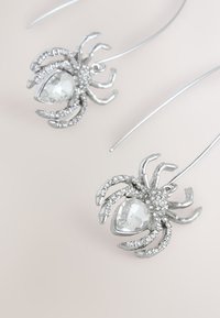 Silver spider-shaped earrings feature a clear faceted stone at the center, surrounded by small rhinestones on the legs and body.