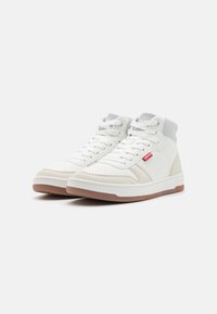 Sneaker alte bianche con un design perforato, accenti grigi e suola in gomma color beige, con un logo rosso sul lato.