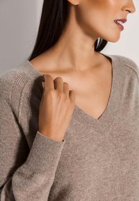 Beige V-Ausschnittpullover aus weichem, strukturiertem Stoff. Mit gerippten Bündchen und Schulterdetails. Nahaufnahme, die Hand und Hals hervorhebt.
