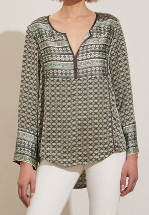 Femme portant un chemisier à manches longues à motifs géométriques verts et bruns, associé à un pantalon blanc, debout devant un fond uni.