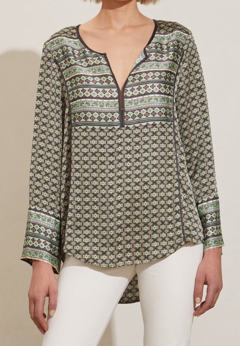 Femme portant un chemisier à manches longues à motifs géométriques verts et bruns, associé à un pantalon blanc, debout devant un fond uni.