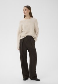 Mujer de cabello largo, vestida con un suéter beige y pantalones negros holgados, de pie con una mano en el bolsillo, mirando hacia la izquierda.