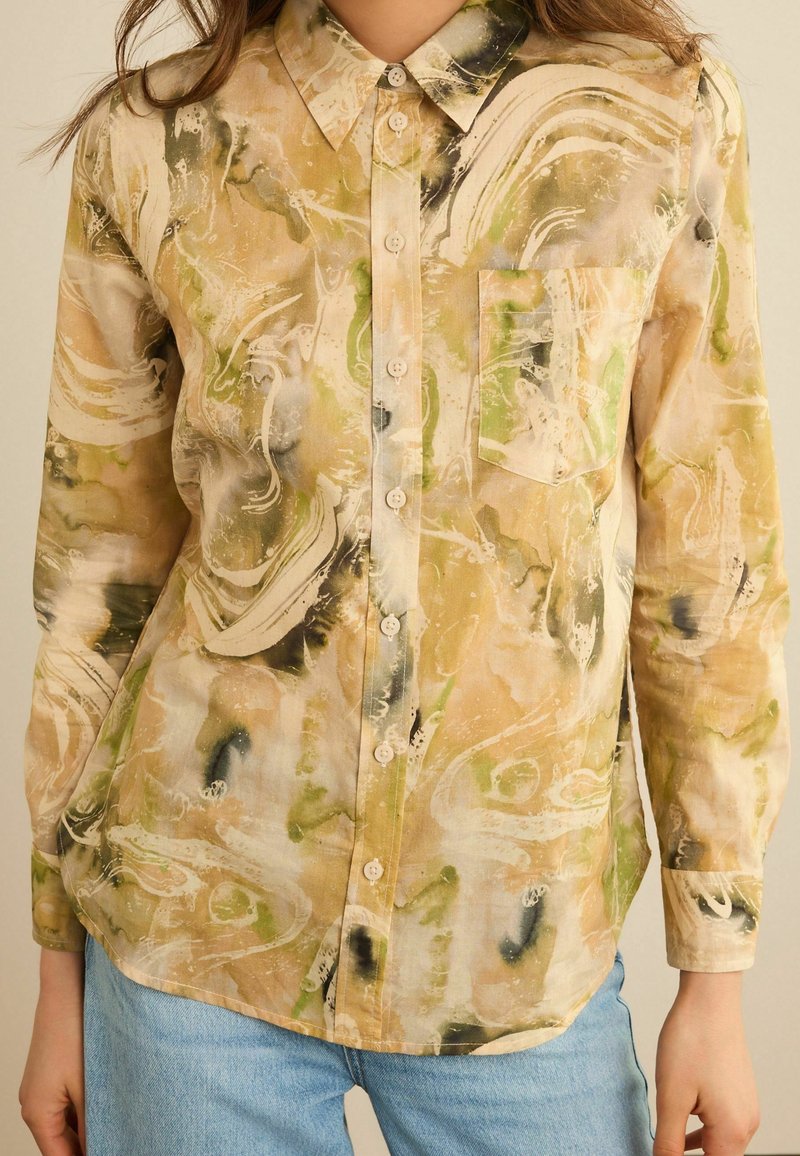 Femme portant une chemise à manches longues beige, verte et noire marbrée avec une poche poitrine et un jean bleu clair.