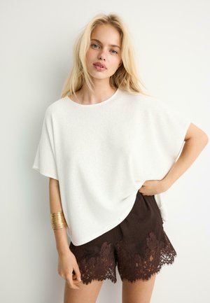 Blonde Frau, die ein locker sitzendes weißes Oberteil und dunkelbraune Shorts mit Spitzenbesatz trägt, steht mit einer Hand in der Hüfte und einem goldenen Manschettenarmband.