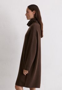 Braun gestricktes Kleid mit hohem Kragen, lockerem Schnitt, langen Ärmeln und gerippten Bündchen, vor einem hellen Hintergrund gezeigt.
