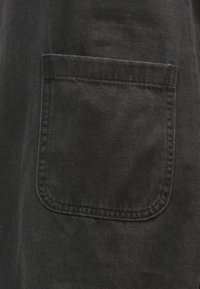 Čierna látka s hladkou textúrou; na pravej strane sa nachádza štvorcová vrecko s prešitými okrajmi, ktoré predstavuje jednoduchý dizajn.