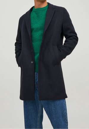Homme portant un manteau bleu marine foncé par-dessus un pull vert, les mains dans les poches du manteau, associé à un jean bleu sur un fond uni.