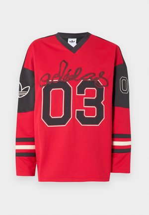 Dlhý športový dres s červenými a čiernymi rukávmi, na ktorom je veľké číslo „03“, logo Adidas na rukávoch a nápis v štýle podpisu cez hruď.