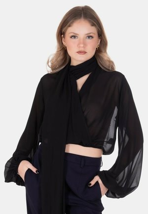 Giovane donna con capelli mossi che indossa una camicetta nera trasparente a maniche lunghe e pantaloni scuri a vita alta, mani nelle tasche, espressione neutra.