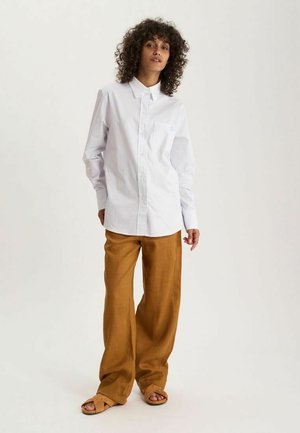 Femme aux cheveux bouclés portant une chemise blanche ample boutonnée, un pantalon large beige et des sandales beiges, debout devant un fond clair uni.