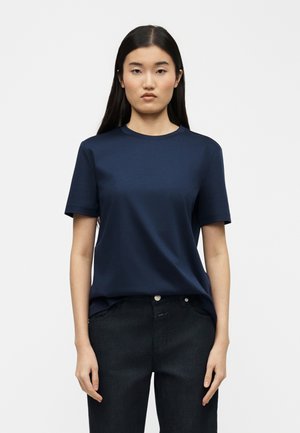 Jeune femme portant un t-shirt à manches courtes uni bleu foncé et un jean noir, debout devant un fond blanc, faisant face à l'avant.