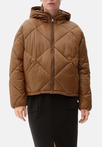 Brązowa kurtka puchowa z kapturem, z pikowanym wzorem w diamenty, z zamkiem z przodu, dopasowana krótka. Noszona na czarnej spódnicy.
