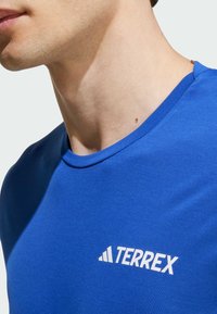 Camisa atlética azul hecha de un tejido suave, con un escote redondo y el logo blanco "TERREX" en el lado izquierdo.