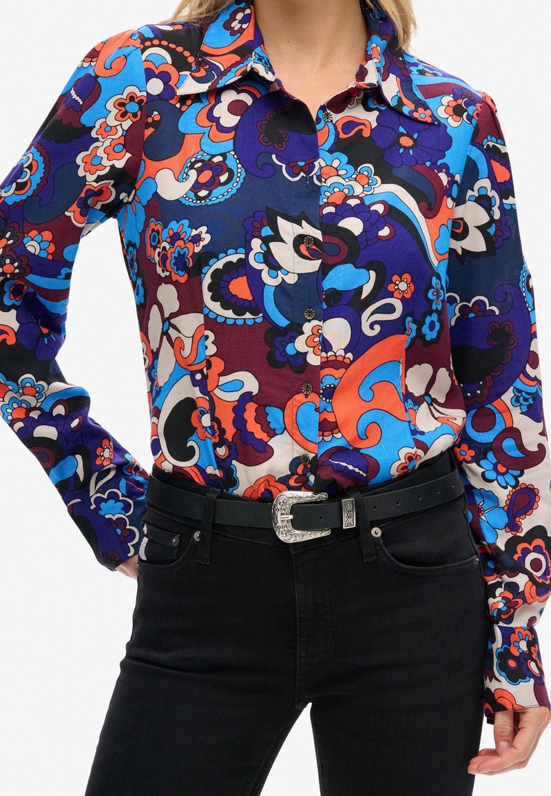 Chemise à boutons multicolore avec un motif floral et cachemire en bleu, orange et violet, associée à un jean noir et une ceinture décorative.