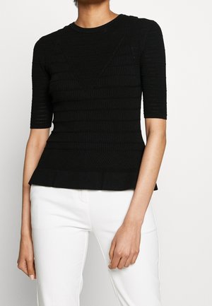 Kobieta ubrana w dopasowaną czarną, teksturowaną bluzkę z krótkim rękawem, w zestawieniu z białymi spodniami na jednolitym tle.