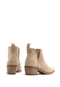 Beżowe zamszowe botki za kostkę z niskim, masywnym obcasem, bocznymi wycięciami i uchwytami ułatwiającymi zakładanie. Gładka struktura i minimalistyczny design.